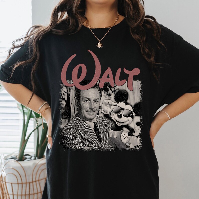 May include: A black t-shirt featuring a graphic print of Walt Disney and Mickey Mouse wearing sunglasses. Above the black and white image, the word "WALT" is printed in a large, dusty rose-colored script font. The design has a distressed texture.