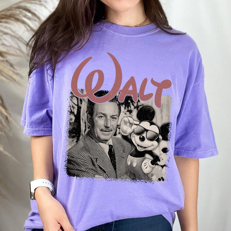 May include: A light purple short-sleeve t-shirt featuring a large graphic print. The design includes the word WALT in a dusty rose script font above a black and white image of Walt Disney standing next to Mickey Mouse wearing sunglasses.
