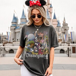 May include: A charcoal gray short-sleeve t-shirt featuring a colorful graphic print of various cartoon characters, a large spherical structure, and a castle. The text "Disneyland Est. 1955" is printed in white lettering at the bottom of the graphic design.
