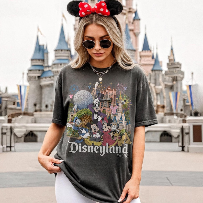 May include: A charcoal gray short-sleeve t-shirt featuring a colorful graphic print of various cartoon characters, a large spherical structure, and a castle. The text "Disneyland Est. 1955" is printed in white lettering at the bottom of the graphic design.