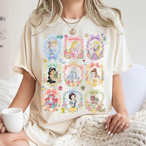 May include: A cream-colored t-shirt featuring a graphic print of nine ornate, pastel-colored frames. Each frame contains a different illustrated character or a castle, surrounded by floral accents and small icons like a lamp, a rose, and a frog.