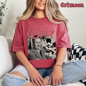May include: A crimson short-sleeve t-shirt featuring a black and white graphic of Walt Disney and Mickey Mouse wearing sunglasses. The word Walt is printed in a large, stylized font above the graphic. The text Crimson appears in the top right.