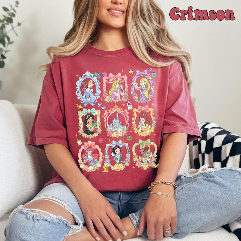 May include: A crimson short-sleeve t-shirt featuring a graphic print of nine ornate, colorful frames. Each frame contains a different illustrated princess or a castle. The text Crimson is printed in the upper right corner in a stylized font.