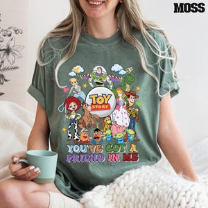 May include: Moss green t-shirt with a vibrant Toy Story character graphic. The design showcases Buzz Lightyear, Woody, and other characters, accompanied by the phrase "YOU'VE GOT A FRIEND IN ME".