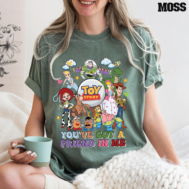 May include: Moss green t-shirt with a vibrant Toy Story character graphic. The design showcases Buzz Lightyear, Woody, and other characters, accompanied by the phrase "YOU'VE GOT A FRIEND IN ME".