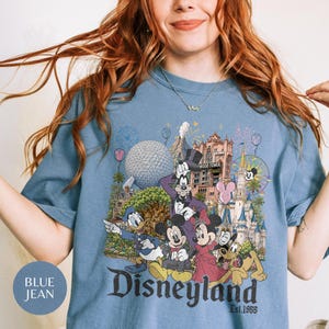 May include: A blue jean colored t-shirt featuring a graphic print of Disney characters including Mickey Mouse, Minnie Mouse, Donald Duck, Goofy, and Pluto in front of a castle and theme park landmarks with the text Disneyland Est. 1955 printed below.