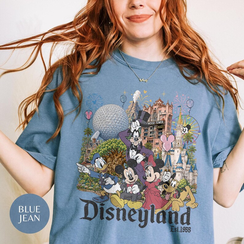 May include: A blue jean colored t-shirt featuring a graphic print of Disney characters including Mickey Mouse, Minnie Mouse, Donald Duck, Goofy, and Pluto in front of a castle and theme park landmarks with the text Disneyland Est. 1955 printed below.