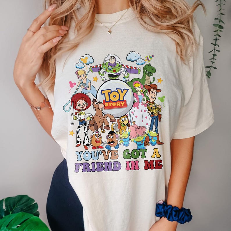 May include: Cream-coloured t-shirt with a vibrant graphic of Toy Story characters, such as Buzz Lightyear, Woody, and Jessie. The text "TOY STORY" is in a circular design, with the phrase "YOU'VE GOT A FRIEND IN ME" underneath.