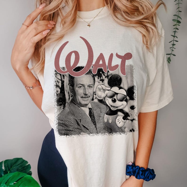 May include: A cream-colored t-shirt featuring a graphic print of Walt Disney and Mickey Mouse wearing sunglasses. The word WALT is printed in large, dusty rose-colored letters above the black and white photographic image of the pair.