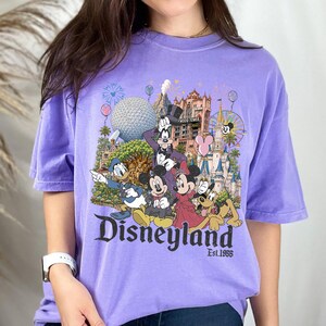 May include: A light purple short-sleeve t-shirt featuring a colorful graphic of Disney characters including Mickey Mouse, Minnie Mouse, Donald Duck, Goofy, and Pluto in front of a castle and theme park landmarks with the text Disneyland Est. 1955.