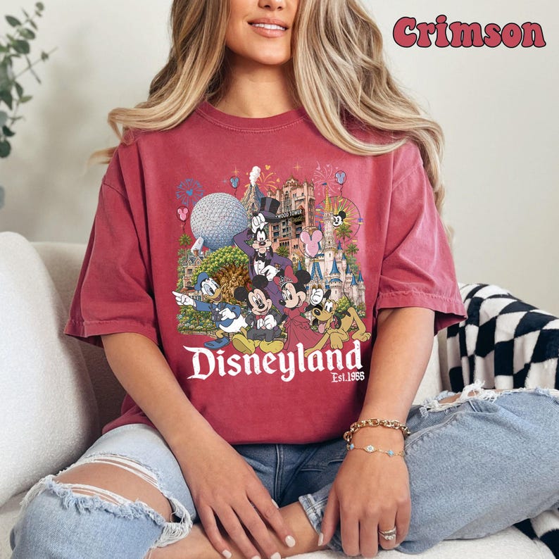 May include: A crimson short-sleeve t-shirt featuring a graphic print of Mickey Mouse, Minnie Mouse, Donald Duck, Goofy, and Pluto in front of a Disneyland castle and Epcot sphere. The text reads Disneyland Est. 1955 and Crimson in the corner.