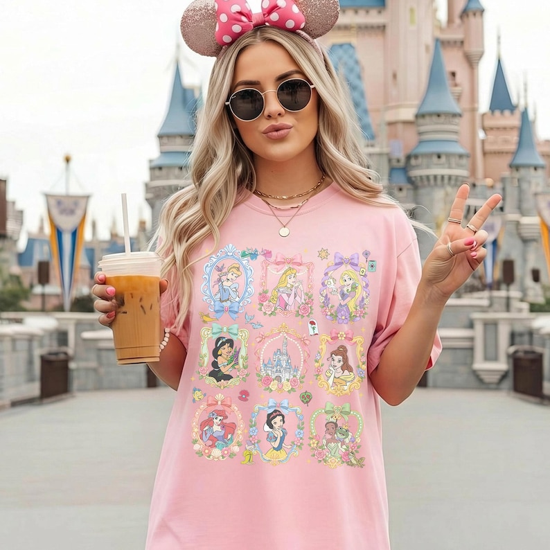 May include: A light pink t-shirt featuring a grid of nine decorative frames, each containing a different illustrated character or castle. The design includes pastel-colored borders, floral accents, and small icons like a rose and a book, printed on the front.