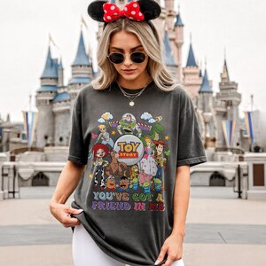 May include: Dark grey t-shirt with a colourful graphic of Toy Story characters. The shirt features the text "Toy Story" and "You've Got a Friend in Me". The t-shirt is crew neck and has short sleeves.