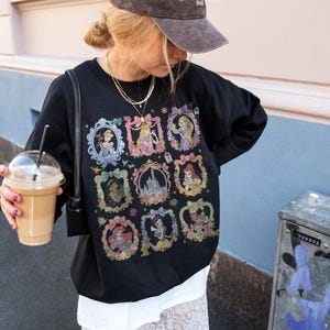 May include: A black long-sleeve sweatshirt featuring a graphic print of nine ornate, colorful frames containing various animated characters and a central castle illustration. The design includes bows, floral accents, and small stars surrounding each frame.