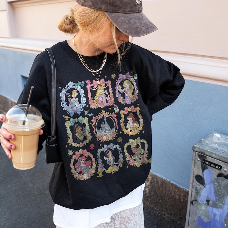 May include: A black long-sleeve sweatshirt featuring a graphic print of nine ornate, colorful frames containing various animated characters and a central castle illustration. The design includes bows, floral accents, and small stars surrounding each frame.