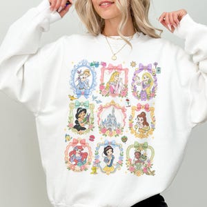 May include: A white long-sleeve crewneck sweatshirt featuring a central graphic print of nine ornate, pastel-colored frames. Inside the frames are illustrations of various animated princesses, a castle, and small icons like a lamp, rose, and frog.