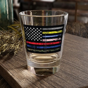Może przedstawiać: Przezroczysty kieliszek do wódki z amerykańską flagą z paskami reprezentującymi różne zawody: korekta, wysyłka, pielęgniarka i strażak. Szklanka stoi na drewnianej powierzchni, z butelką i suszonymi roślinami w tle.
