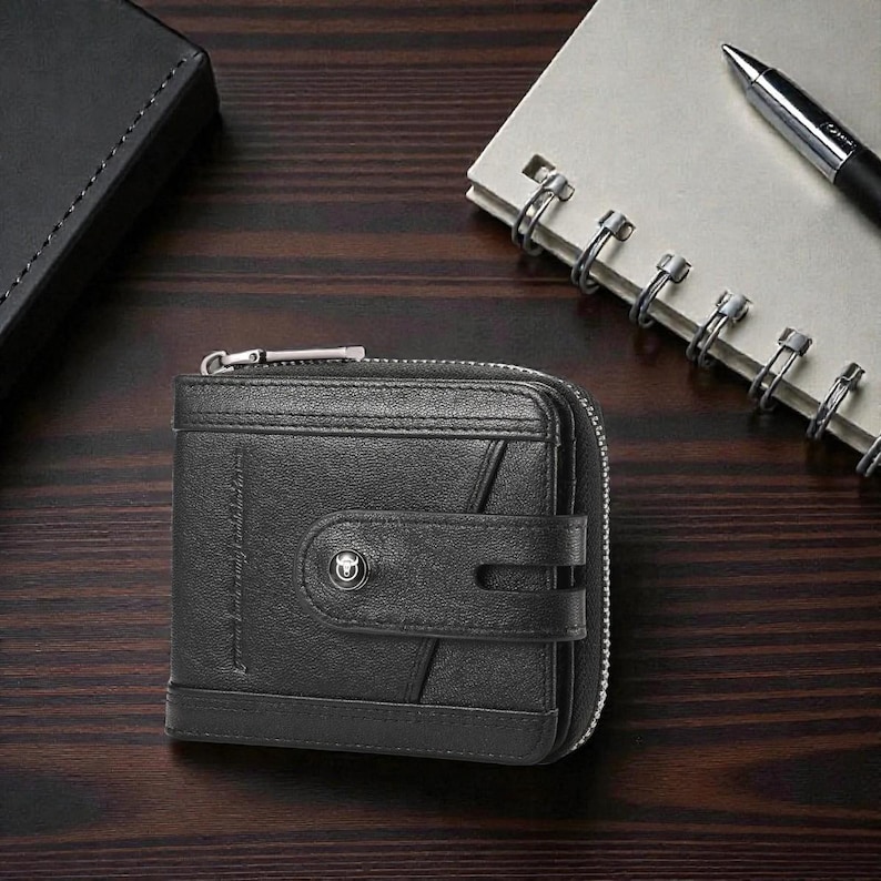 May include: Black leather wallet with a zipper closure and a snap closure. The wallet has a small logo on the front and is sitting on a wooden surface. A notebook and pen are in the background.