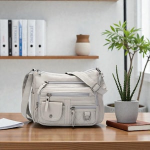 May include: A light gray crossbody bag with multiple zippered compartments and a shoulder strap. The bag has a front flap pocket with a snap closure and a decorative stitched pattern. The bag is sitting on a wooden surface.