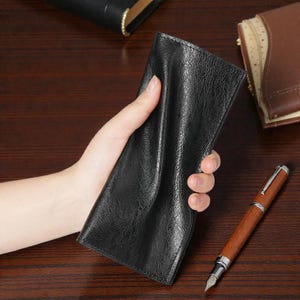 May include: A black leather wallet with a textured finish is held in a hand. A wooden pen with silver detailing lies on a dark wooden surface, alongside other leather items.
