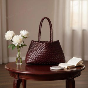 Puede incluir: Un bolso tote de color burdeos oscuro con una construcción de cuero tejido y dos asas trenzadas a juego. El bolso tiene una base estructurada y una abertura ancha, mostrando un patrón de cuadrícula consistente en toda la superficie exterior.