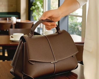 May include: A brown leather handbag with a structured design. The bag features a top handle, a flap closure, and white stitching details. The bag is held by a person in a white outfit.