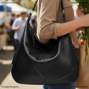 May include: A black hobo bag with a smooth, textured surface, a single shoulder strap, and a vertical seam detail running down the centre. The bag features silver-toned metal hardware rings connecting the strap to the main body.