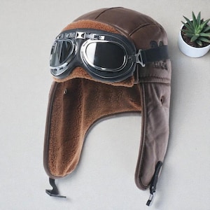 Puede incluir: Gorro de aviador marrón con gafas incorporadas. El gorro tiene un forro de piel sintética y orejeras. Las gafas tienen montura plateada y lentes oscuras. El gorro es de material similar al cuero y tiene una estética vintage.
