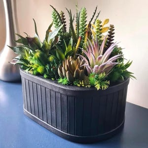 May include: An oval black planter filled with artificial succulents in shades of green, yellow, and purple. The planter has a ribbed design. The arrangement is a decorative piece.