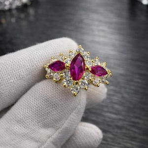 Puede incluir: Anillo dorado con una piedra central magenta en forma de óvalo, flanqueada por dos piedras similares más pequeñas. El anillo está adornado con numerosas piedras pequeñas y transparentes.
