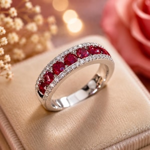 May include: A silver-toned band ring featuring a central row of five round-cut deep red rubies. The rubies are flanked on both the top and bottom edges by two parallel rows of small, clear, round-cut white stones set into the metal band.