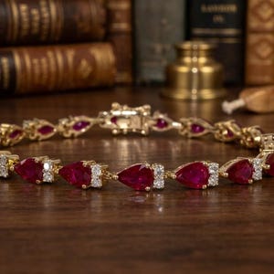 Könnte beinhalten: Ein goldenes Armband mit birnenförmigen, tiefroten Edelsteinen und kleinen weißen Diamanten. Das Armband liegt auf einer dunklen Holzoberfläche, mit antiken Büchern im Hintergrund. Der Verschluss ist sichtbar.