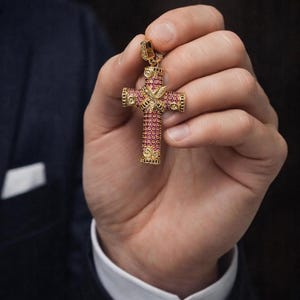 May include: A gold-colored cross pendant with a detailed design. The cross is adorned with small, pink gemstones and features a bird emblem at the center. The pendant is held up by a hand, showcasing its intricate craftsmanship and luxurious appearance.