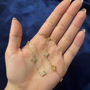 Puede incluir: Un delicado collar de oro con colgantes en forma de trébol. Algunos tréboles están adornados con pequeñas y brillantes piedras preciosas. El collar se muestra sobre un fondo azul oscuro, resaltando el intrincado diseño y brillo de la joya.