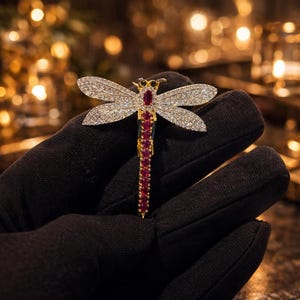 Puede incluir: Broche de libélula con cuerpo dorado, detalles en color rubí y adornos similares a diamantes en las alas. El broche se sostiene sobre un fondo borroso, mostrando su intrincado diseño y detalles brillantes.