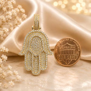 May include: A gold-toned Hamsa hand pendant completely encrusted with small clear stones, featuring a matching stone-set bail at the top. The pendant is positioned next to a United States one-cent copper coin for scale, showing the relative size.