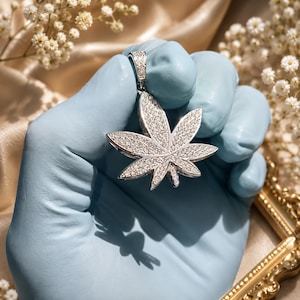 May include: A silver-toned pendant shaped like a seven-pointed cannabis leaf, fully encrusted with small, clear, sparkling stones. The pendant features a matching silver-toned bail at the top, also set with clear stones, designed for attachment to a chain.