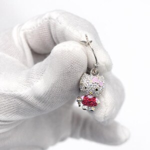 Puede incluir: Un pendiente plateado con un diseño de Hello Kitty. El dije está incrustado con cristales transparentes y rojos, con un lazo y detalles rosas. El pendiente es sostenido por una mano enguantada sobre un fondo blanco.