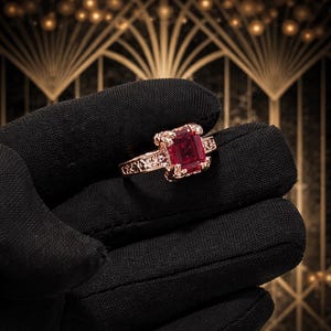 Könnte beinhalten: Ein roségoldener Ring mit einem großen, quadratisch geschliffenen, roten Edelstein. Der Ring wird von einer behandschuhten Hand gehalten, die das aufwendige Filigranmuster auf dem Band und die leuchtende Farbe des Edelsteins zeigt. Der Hintergrund hat ein goldenes Art-Deco-Design.