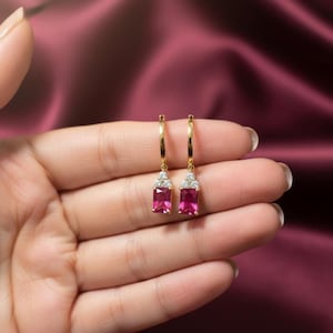 Puede incluir: Un par de pendientes de aro dorados con piedras preciosas rectangulares de color magenta. Cada pendiente presenta un grupo de pequeñas piedras preciosas transparentes encima de la piedra magenta. Fondo burdeos.