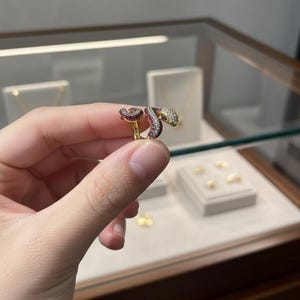 May include: A gold-colored snake ring with a detailed design. The snake's head and tail are adorned with clear and red gemstones. The ring is held in a hand, showcasing its intricate craftsmanship and elegant style.