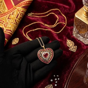 May include: A gold necklace with a heart-shaped pendant. The pendant features a central red gemstone surrounded by white stones and an ornate gold filigree design. The necklace is displayed on a red velvet surface.