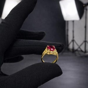 May include: A gold ring featuring a large, square-cut red gemstone, possibly a ruby, set in an ornate, filigree design. Small, clear accent stones are set around the main stone. The ring is held by a gloved hand against a dark background.
