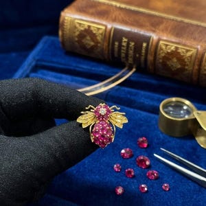 Puede incluir: Broche dorada con forma de abeja, adornada con rubíes y diamantes. El cuerpo de la abeja está formado por numerosos rubíes pequeños, con alas grabadas. Varios rubíes sueltos están dispersos sobre una superficie de terciopelo azul.