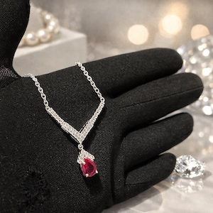 May include: A silver necklace with a V-shaped pendant. The pendant features a teardrop-shaped, red gemstone and is accented with small, clear stones. The necklace is displayed on a black surface, with a diamond and a pearl necklace in the background.