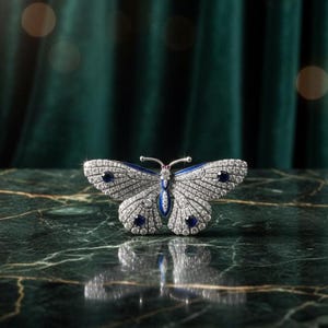 Op de afbeelding: Een vlinderbroche met een zilverkleurig metalen frame, bezet met heldere kristallen. De vleugels hebben blauwe accenten en donkerblauwe edelstenen. Een kleine rode edelsteen is in het midden van het lichaam gezet. De broche wordt getoond op een groen marmeren oppervlak.