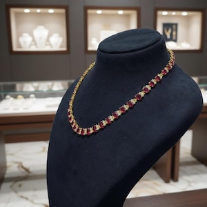 Può includere: Una collana d'oro con pietre preziose rosse taglio ovale e piccole pietre bianche. La collana è esposta su un busto di gioielli nero. Lo sfondo presenta vetrine con altri gioielli.