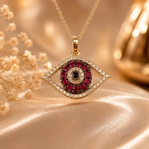 Puede incluir: Un collar de oro con un colgante en forma de ojo. El colgante está incrustado con pequeños diamantes blancos en el borde exterior, un anillo de rubíes de color rojo intenso en el centro y una piedra negra en el medio del diseño del ojo.