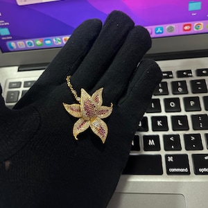 Könnte beinhalten: Eine goldfarbene Anhängerhalskette mit einem Blumenmuster. Die Blume hat rote und klare Edelsteinakzente. Die Halskette wird auf einer schwarzen Oberfläche mit einer Laptop-Tastatur im Hintergrund präsentiert.