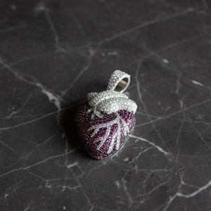 May include: A heart-shaped pendant with a silver bail. The pendant is encrusted with small, sparkling, deep purple and clear stones. The veins of the heart are outlined in clear stones. The pendant is on a dark, marbled surface.